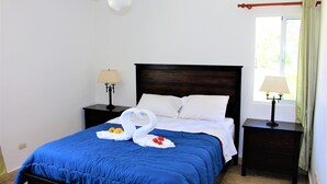 A neatly made bed with a blue bedspread, two white pillows, and a decorative swan made of towels. There are two bedside tables with lamps, a ceiling fan, and a window with curtains. - CORTE SEA Luxury apartment in the heart of Bavaro (Punta Cana)