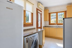A kitchen with a washing machine, a refrigerator, wooden cabinets, and a window with a view of trees.
