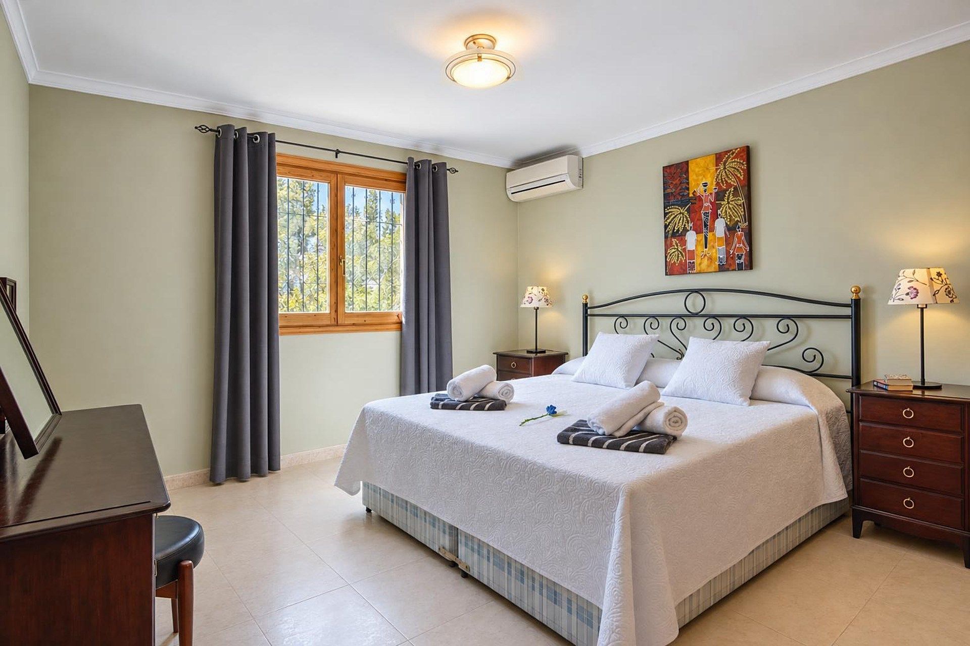 A hotel room with a bed, a desk, a chair, a window with curtains, an air conditioning unit, a painting on the wall, and a lamp on the bedside table.