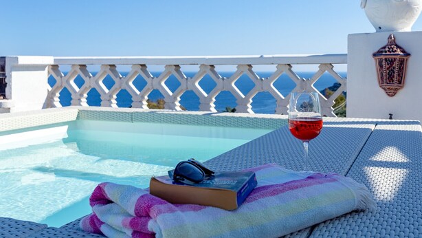 A pool area with a glass table, a book, and a glass of red wine.