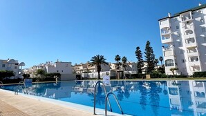 Une piscine à l’eau d’un bleu cristallin, entourée de bâtiments blancs dotés de balcons, sous un ciel dégagé.