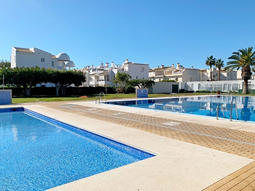 A swimming pool area with clear blue water, adjacent to a row of white buildings with flat roofs and a palm tree in the foreground.