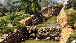 Un élément décoratif aquatique bordé de pierres, avec une petite cascade, entouré d’une végétation luxuriante et, au fond, un bâtiment blanc doté d’un toit recouvert de tuiles.