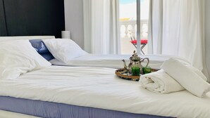 Un lit bien fait, avec du linge de lit blanc et un coussin bleu, un plateau avec une théière et des verres, et un balcon avec des rideaux blancs et une chaise rouge.