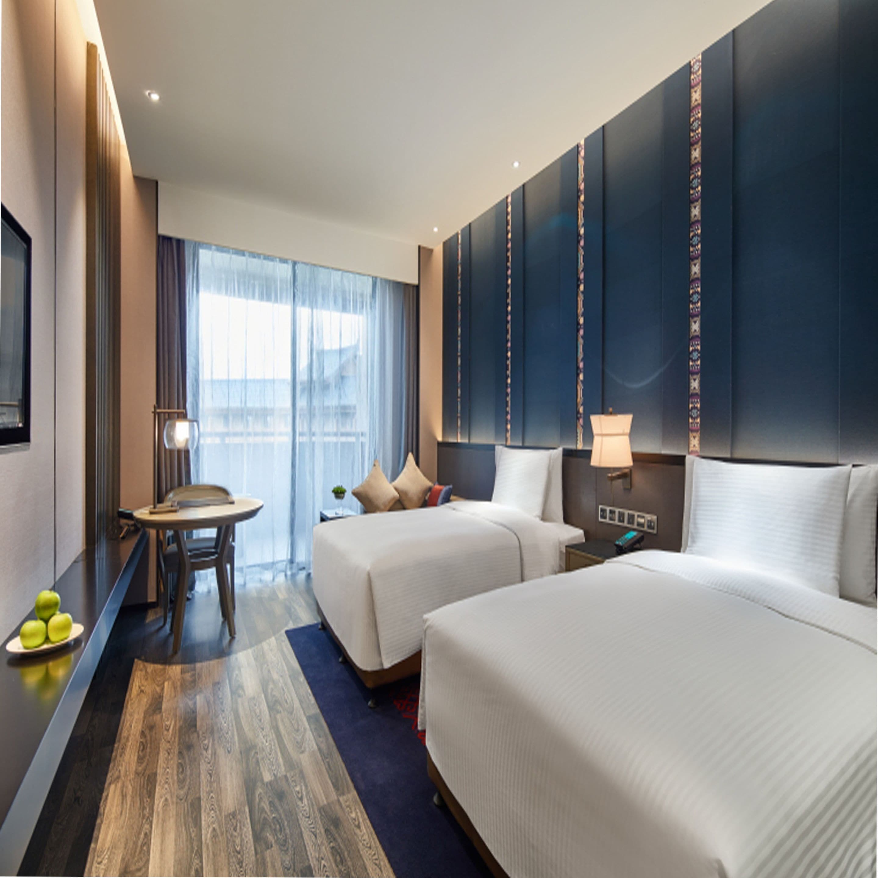 A hotel room with two beds, a desk, a chair, a lamp, and a view of the city through large windows.