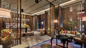 Un moderno vestíbulo de hotel con una decoración de inspiración asiática tradicional, que presenta una zona de estar central con sofás y una mesa de centro, rodeada por separadores decorativos y una alfombra estampada.