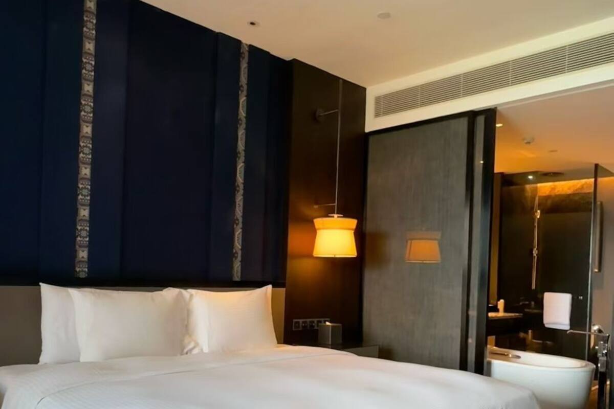 Una cama grande con ropa blanca, un cabecero azul oscuro, una lámpara colgante y un baño visible con bañera.