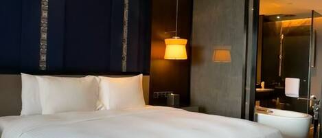 A large bed with white linens, a dark blue accent wall, a hanging pendant light, and a visible bathroom with a bathtub.