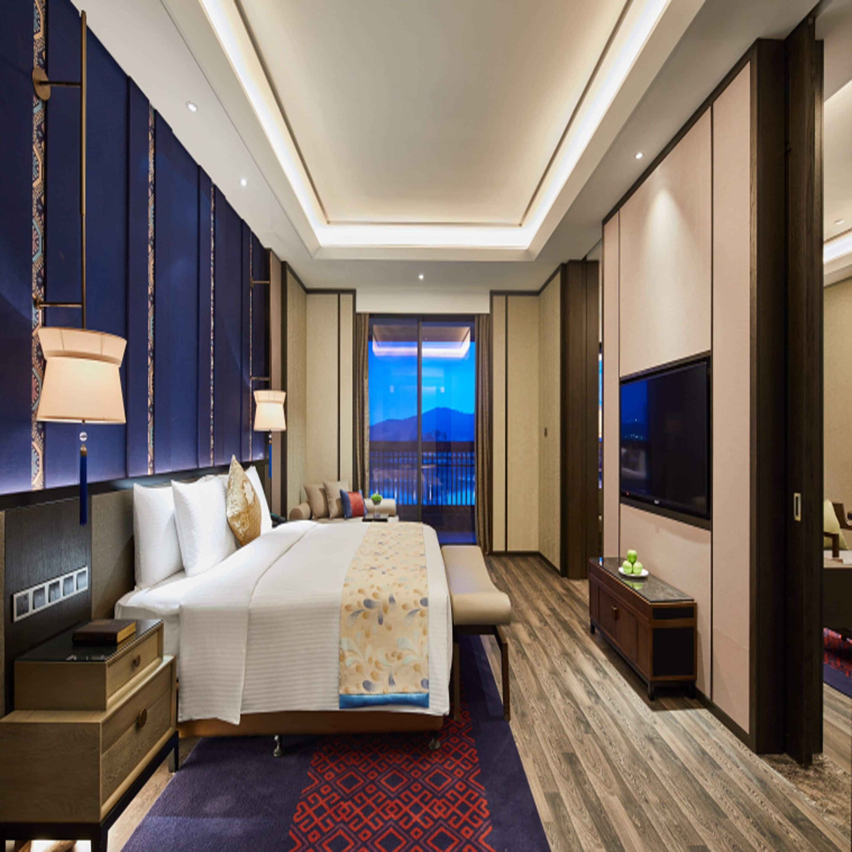 Habitación de hotel moderna con una cama grande, mesitas de noche, televisión y un balcón con vistas a la ciudad.