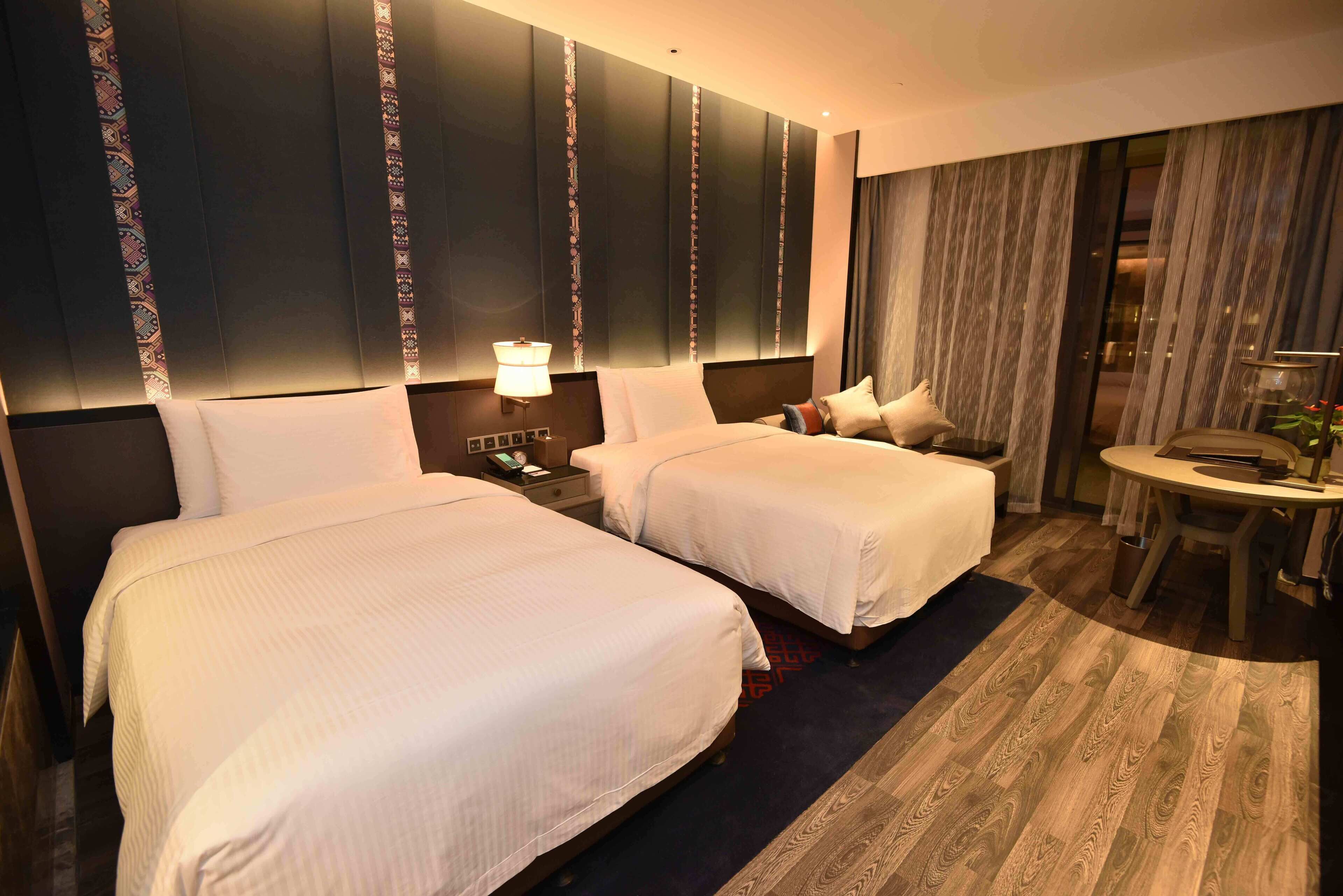 A hotel room with two beds, a small table, a chair, and a nightstand with a lamp.