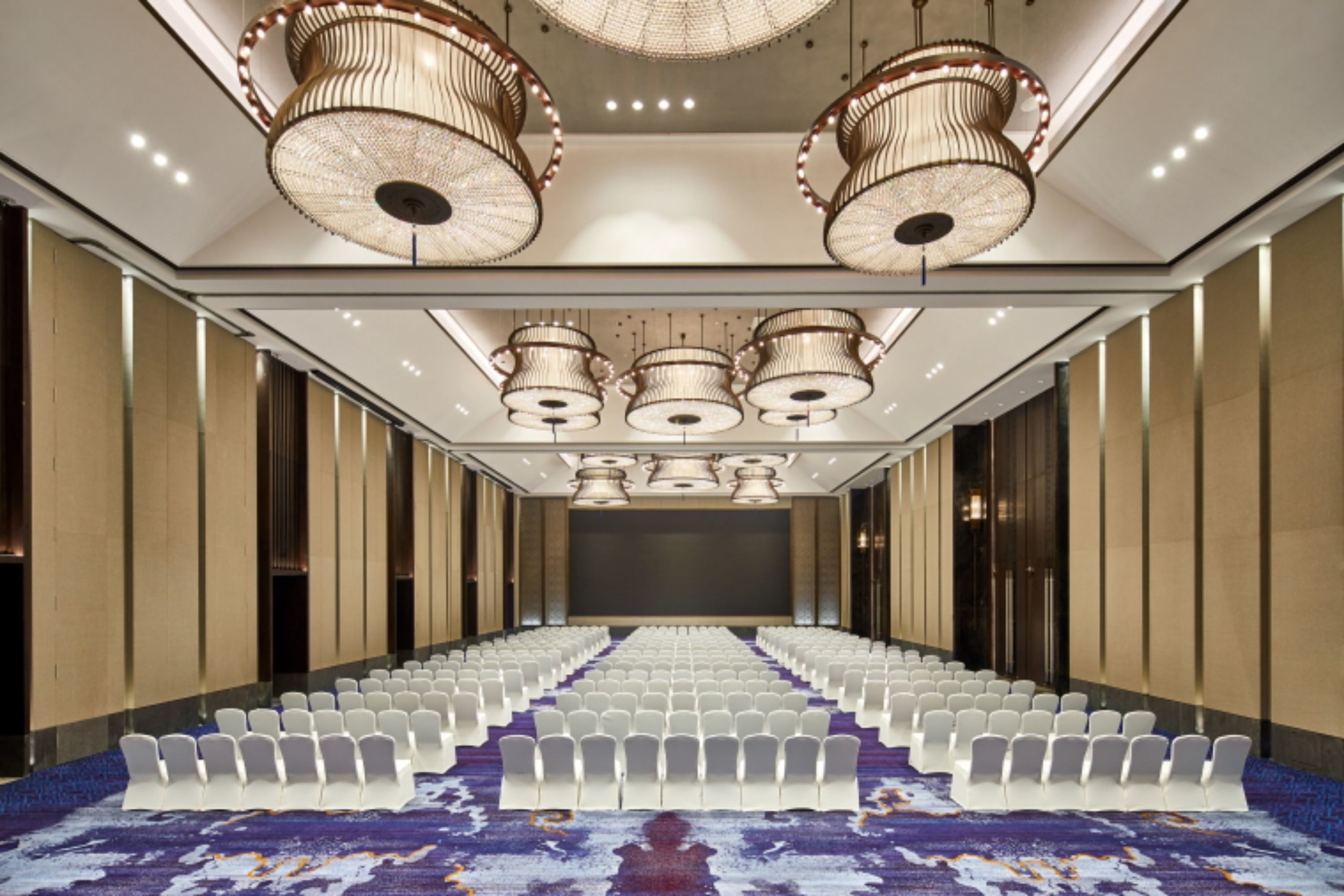 A conference room with rows of white chairs, a large screen at the front, and ornate chandeliers.