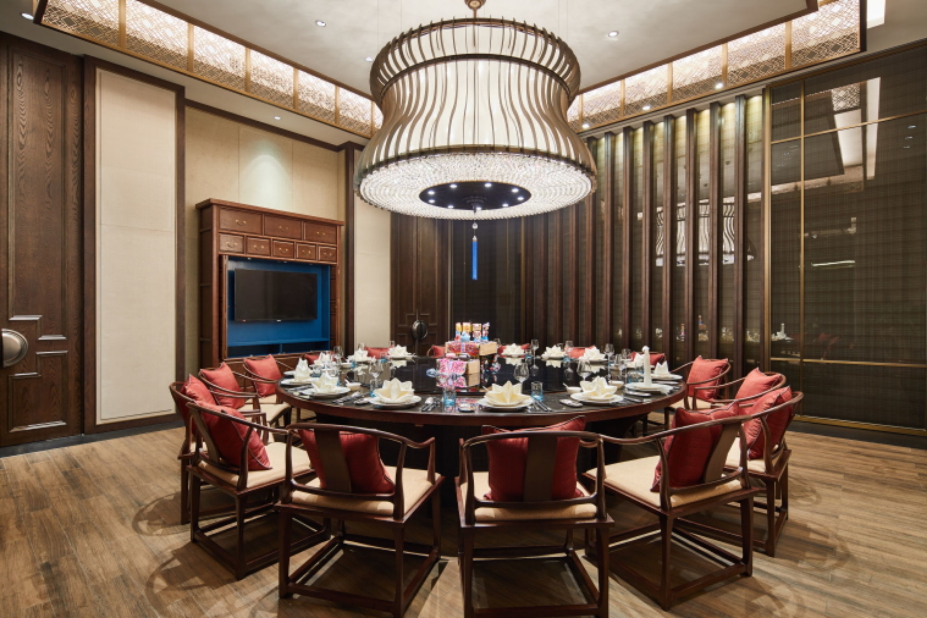Une salle à manger élégante, avec une table ronde dressée pour huit personnes, entourée de chaises à revêtement rouge. Un grand lustre somptueux est suspendu au plafond, et un téléviseur est fixé au mur, à gauche.