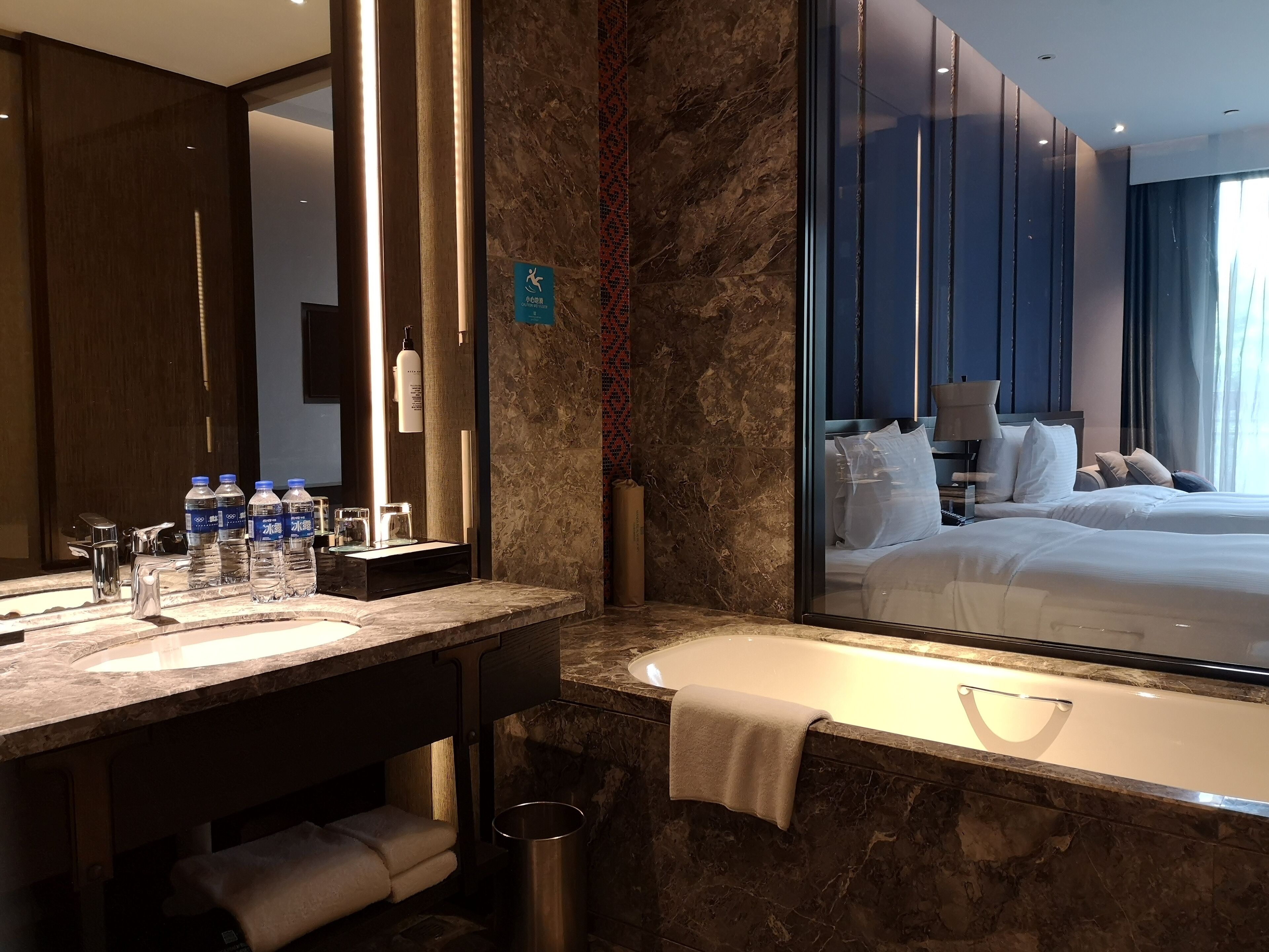 Une salle de bain d’hôtel moderne, dotée d’un grand miroir, de deux vasques, d’une baignoire et d’un grand lit visible à travers une paroi en verre.