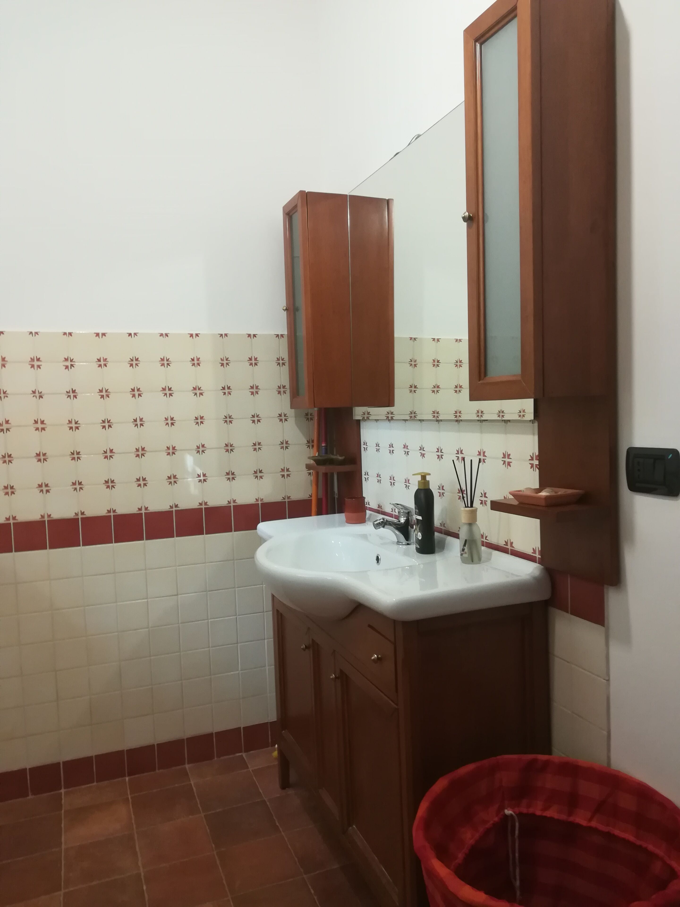 Casa de banho com uma pia branca, armários de madeira, um espelho e um balde vermelho.
