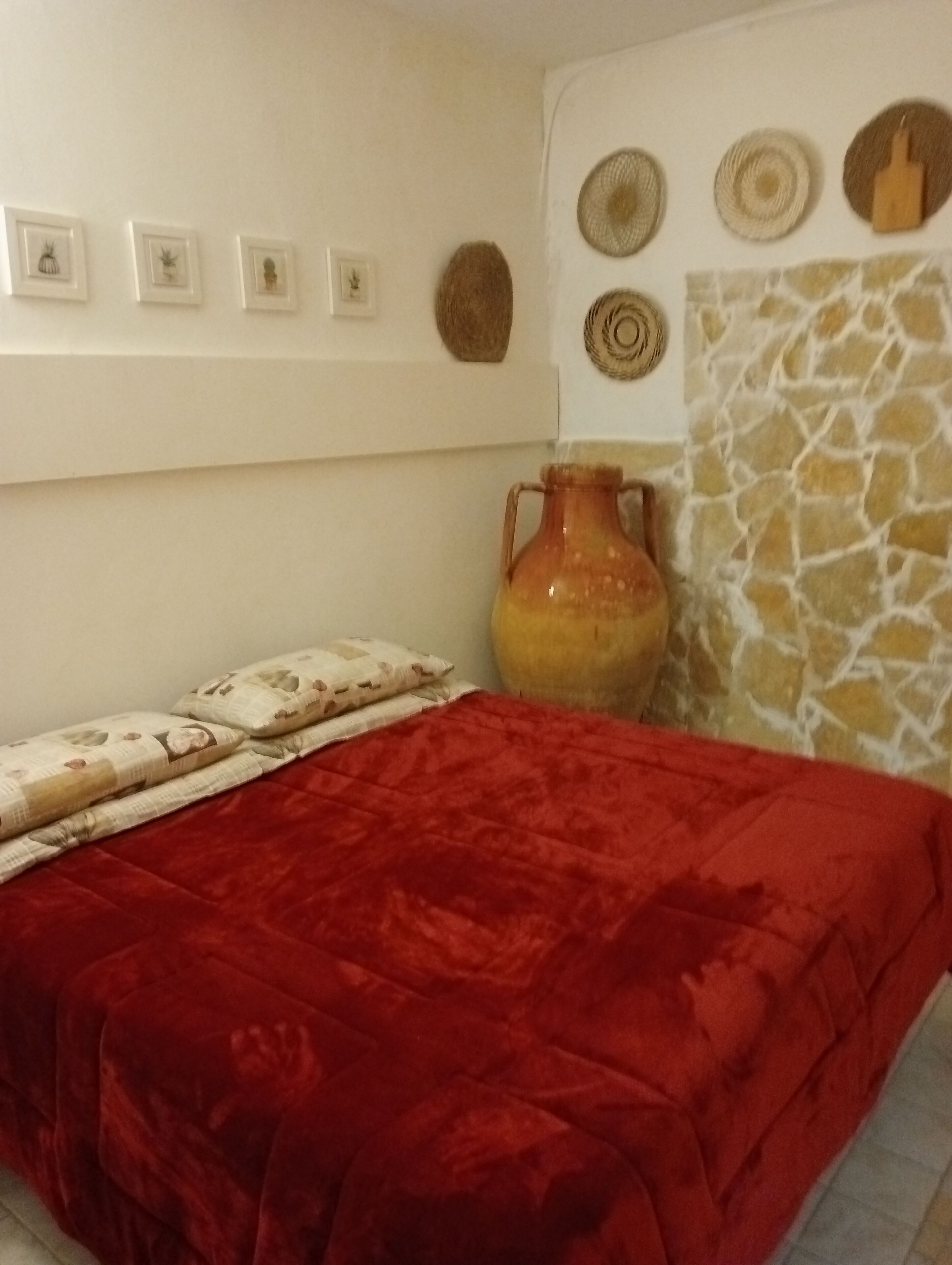 A bedroom with a red bedspread, a large vase, and wall decorations including framed pictures and woven items.