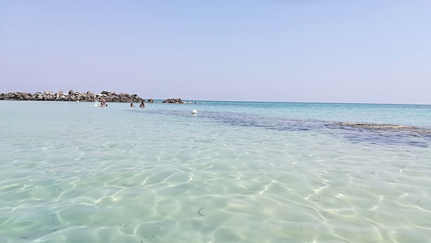 Een helder strand met ondiep water, rotsen op de achtergrond en enkele mensen in de verte.