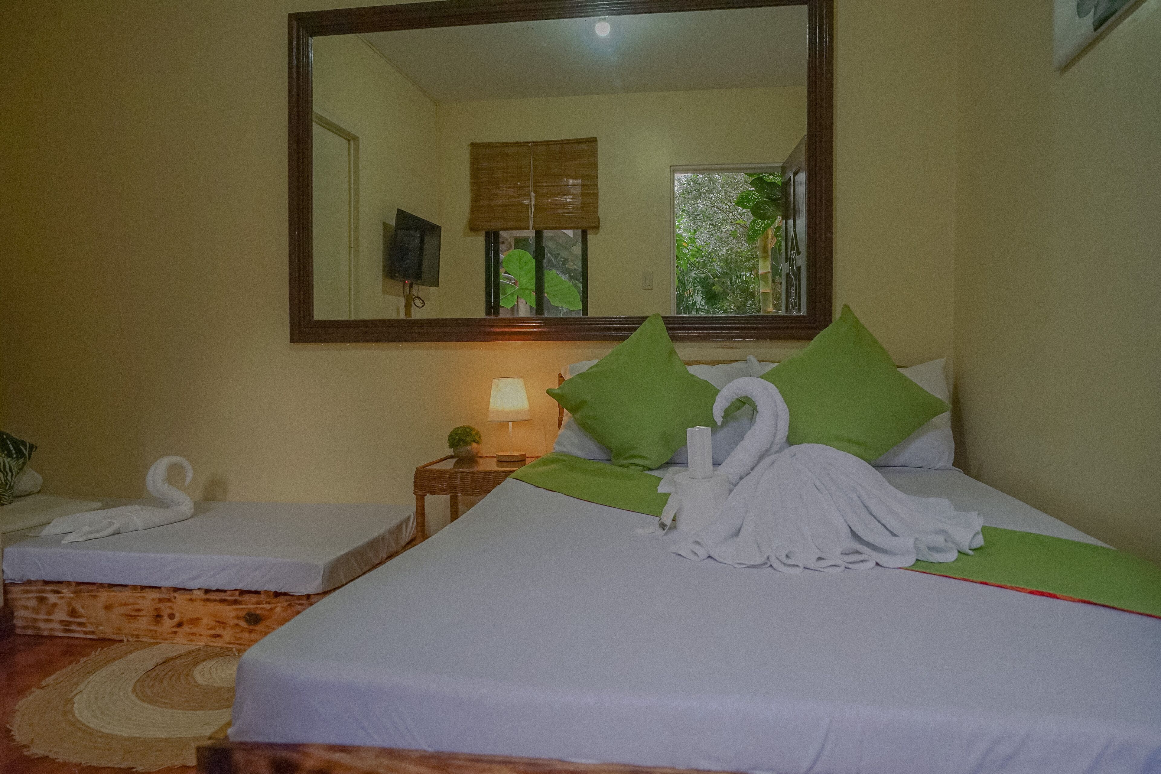 Habitación de hotel con dos camas, un espejo grande, una mesita con lámpara y vistas a zonas verdes.