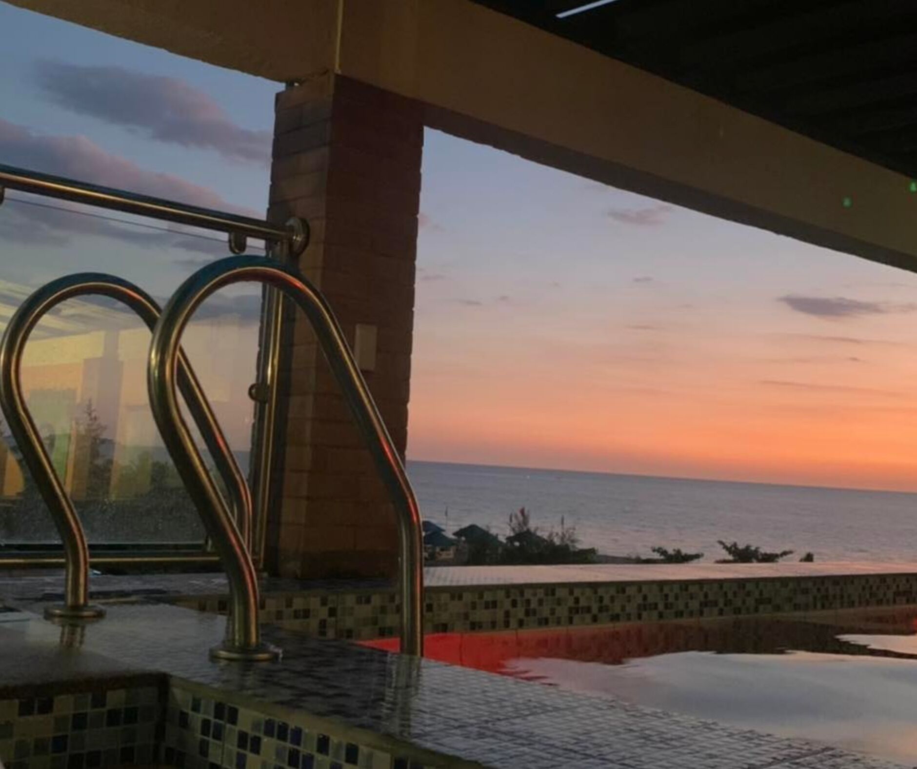 Un área de piscina con vistas a la puesta de sol sobre el océano, una escalera de metal y suelo de baldosas.