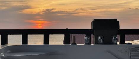 A sunset view from a balcony with a railing, a small seating area, and a view of the sea.