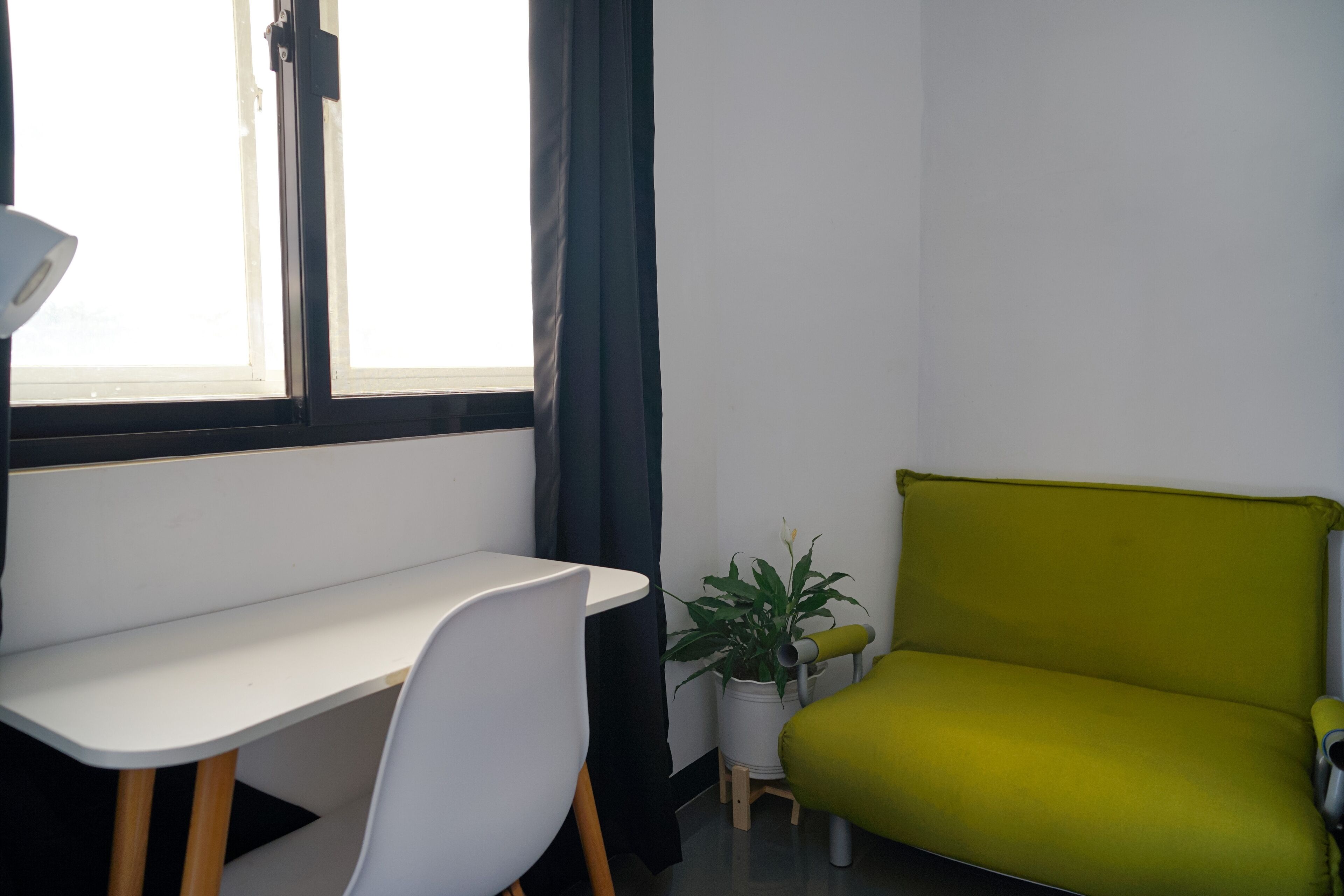 Una habitación con un escritorio blanco, una silla verde, una planta en maceta y un ventanal con cortinas.