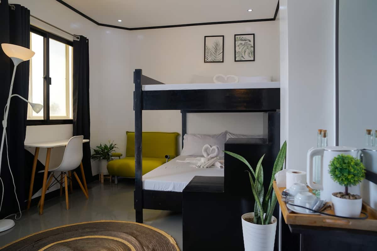 Una habitación de hotel moderna con una cama nido, un escritorio con una silla, un pequeño sofá y una mesita de noche con una lámpara y una planta.