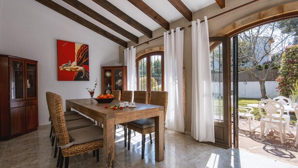 A dining area with a wooden table and wicker chairs, a painting on the wall, and a view through a glass door to an outdoor patio.