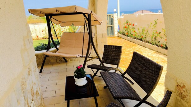 Un espace patio aménagé avec un fauteuil suspendu, deux chaises en osier, une petite table ornée d’un pot de fleurs, et une vue sur la mer.
