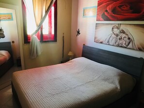 A neatly made bed with a quilted cover, a bedside lamp, a wall with a framed picture, and a window with curtains.