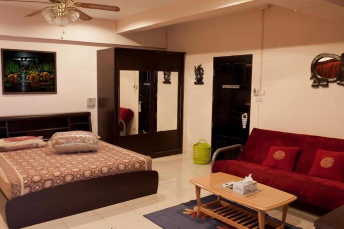 A bedroom with a bed, a red sofa, a wooden coffee table, a ceiling fan, and a painting on the wall.