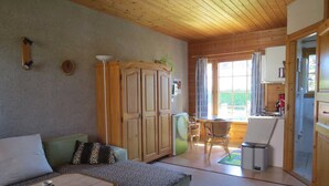 A cozy living room with a sofa, a small table with a decorative item, and a wooden wardrobe. There is a dining area with a table and chairs, and a kitchen visible through an open door. - Wockninsee - Finnish log house & round log house (Ückeritz)