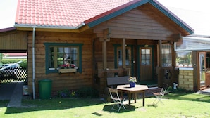 A wooden cabin with a red-tiled roof, a porch with a bench, and a small outdoor seating area with a table and chairs. - Wockninsee - Finnish log house & round log house (Ückeritz)