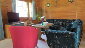 A wooden cabin interior with a red chair, a patterned sofa, a wooden table, a television, and a window with curtains.