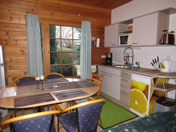 A wooden cabin interior with a dining table, chairs, a kitchen with a sink and appliances, and a view through a large window. - Baltic Sea - Finnish log house & round log house (Ückeritz)