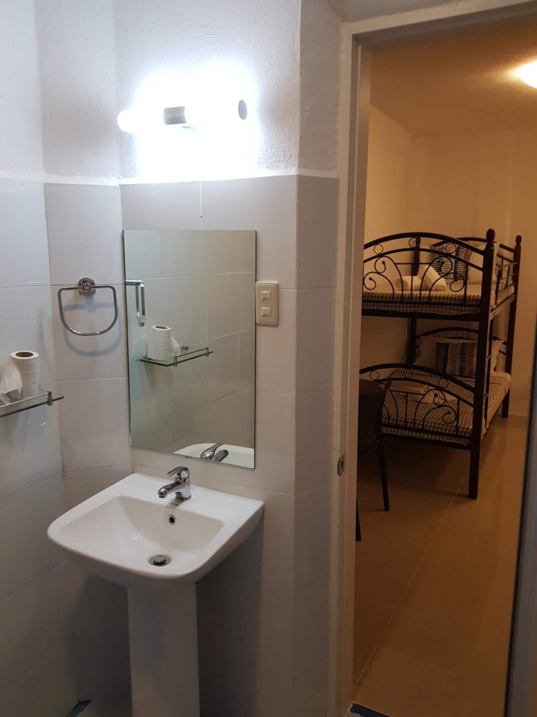 Baño con lavamanos blanco, espejo y zona de ducha. Se ve una cama litera al fondo.