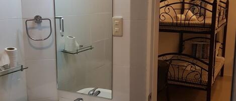 Baño con lavamanos blanco, espejo y zona de ducha. Se ve una cama litera al fondo.