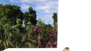 A person sitting on a blue cushioned bench outside a white building with a view of trees and pink flowers. - D' Gecko Hotel (Moalboal)