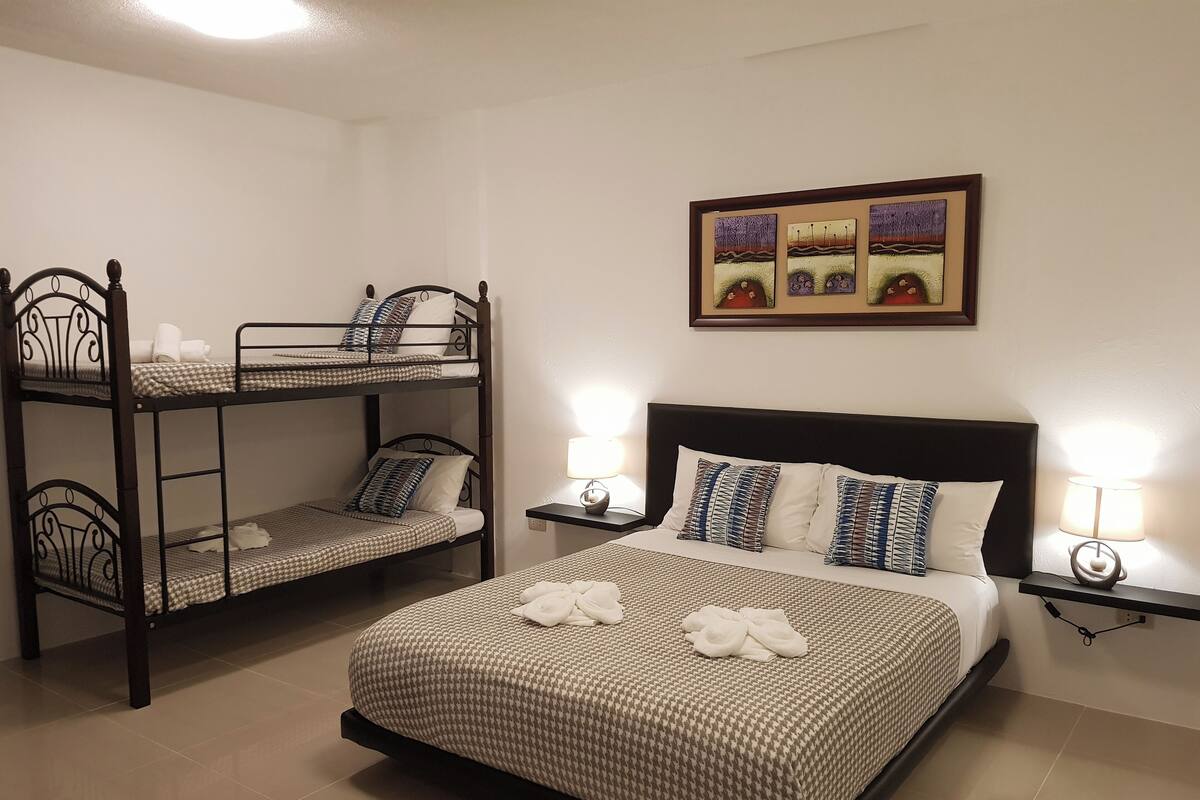 A room with a bunk bed, a single bed, and a nightstand with a lamp. There is a framed picture on the wall above the single bed.
