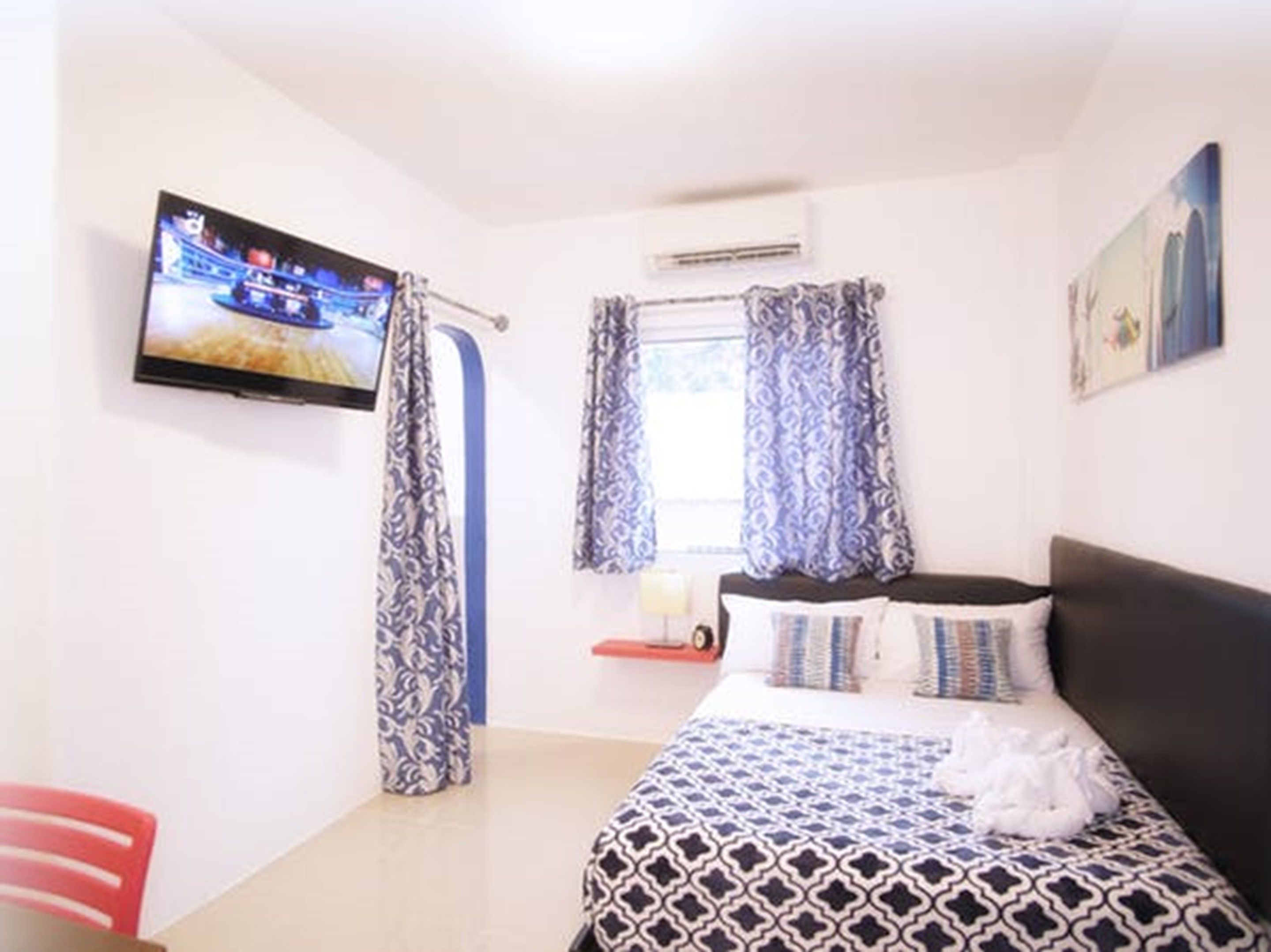 A bedroom with a bed, a TV, a window with curtains, and a painting on the wall.