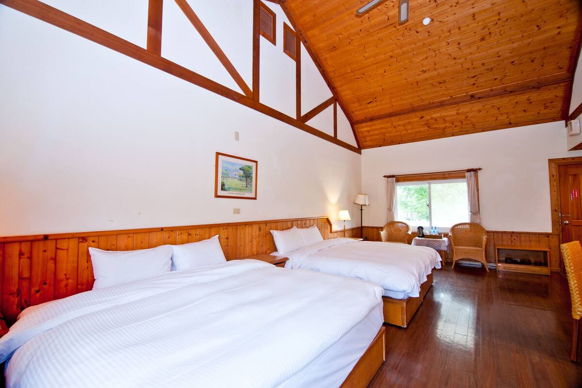 A room with two beds, wooden beams on the ceiling, a painting on the wall, and a window with curtains.