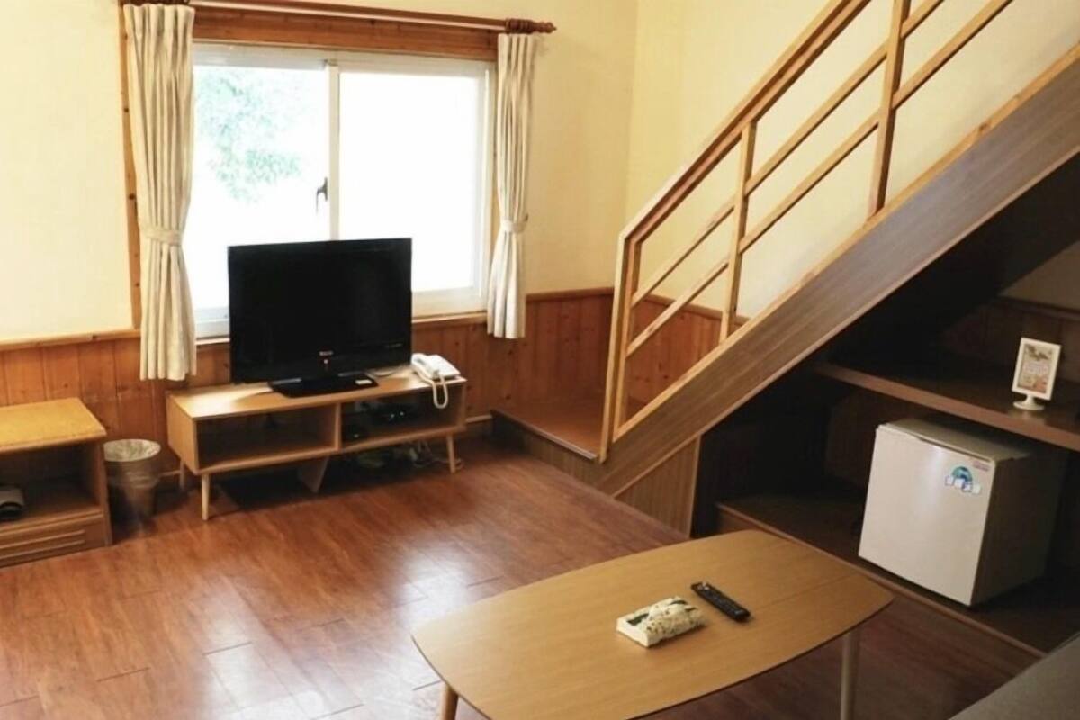 A small, cozy room with a wooden floor, a television on a wooden stand, a small table with a remote control, a refrigerator, and a staircase.