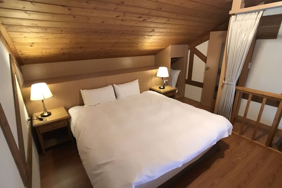 A bedroom with a large bed, two bedside tables with lamps, wooden ceiling, and a wooden railing.