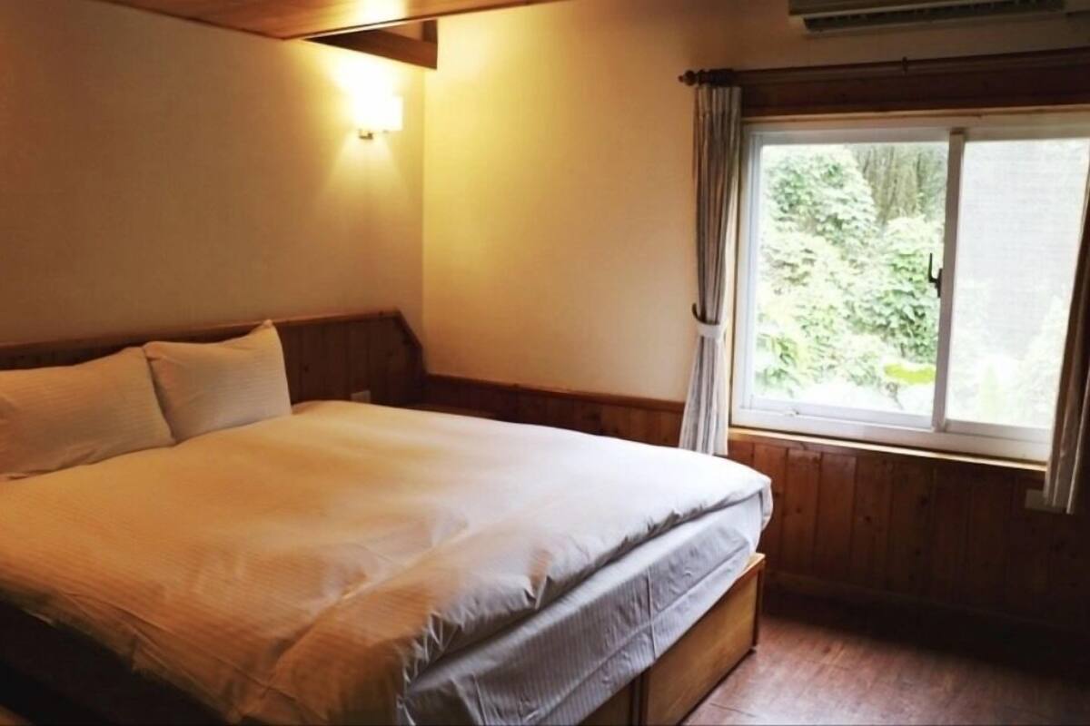 A bedroom with a large bed, wooden headboard, a window with curtains, and a wall-mounted light fixture.