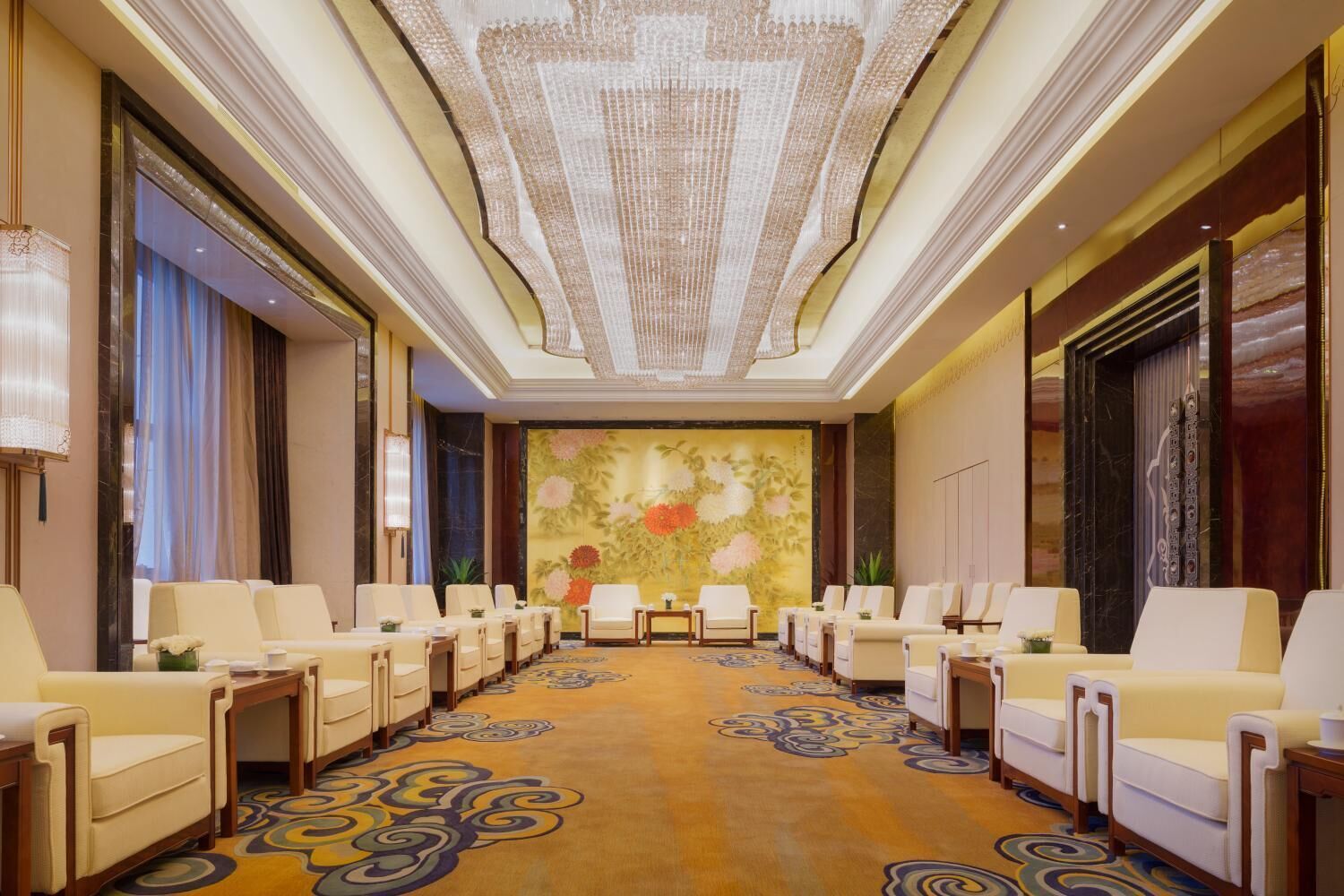 Un hall de hotel con una larga fila de sillas, una alfombra estampada y un gran mural floral en la pared.