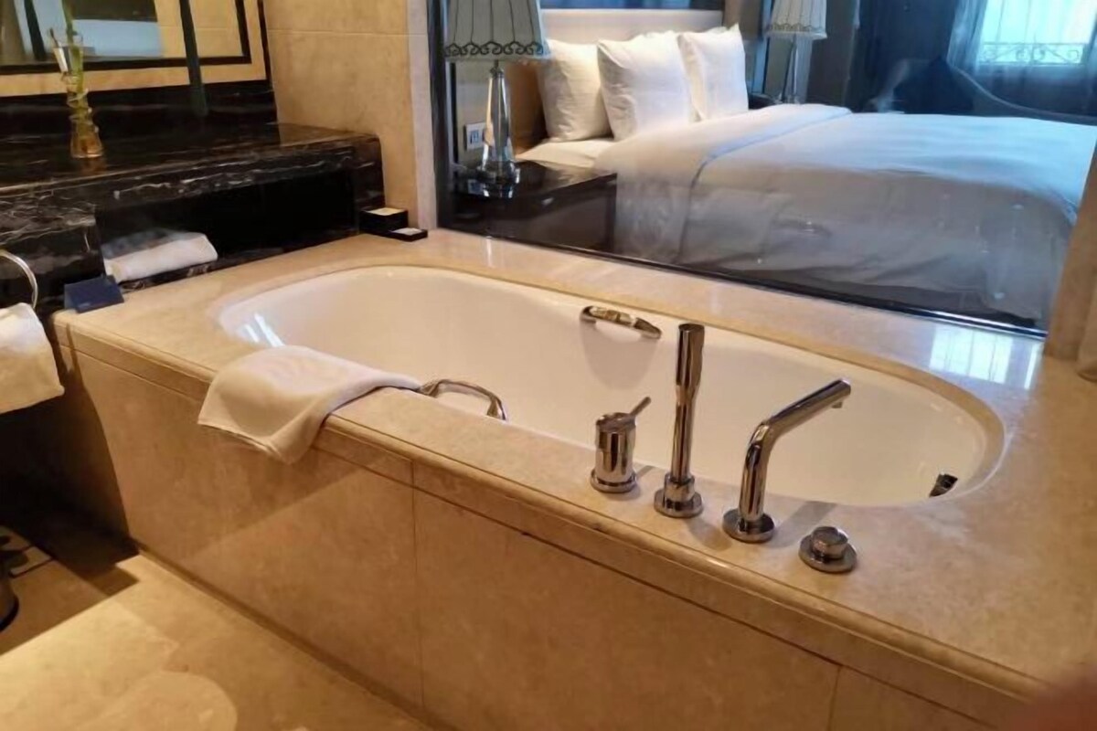 A modern bathroom with a large bathtub, a vanity with a mirror, and a bed visible through the glass partition.