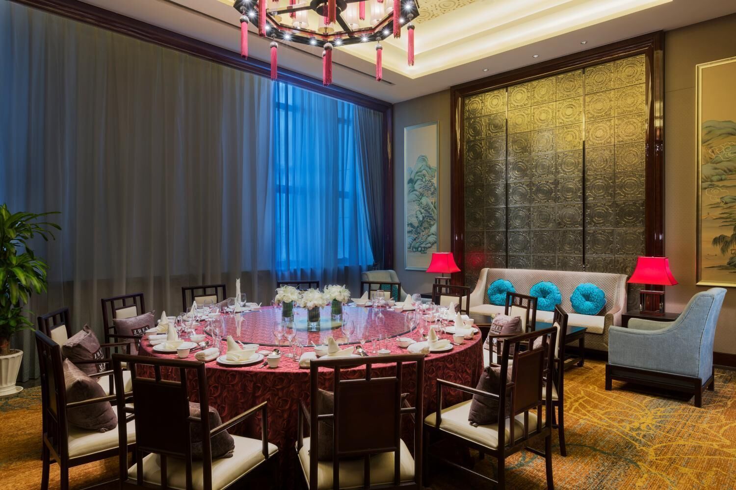 Une salle à manger élégante, avec une table ronde dressée pour six personnes, entourée de chaises, dans une pièce dotée d’une grande fenêtre, d’un canapé avec des coussins bleus et d’une lampe rouge.