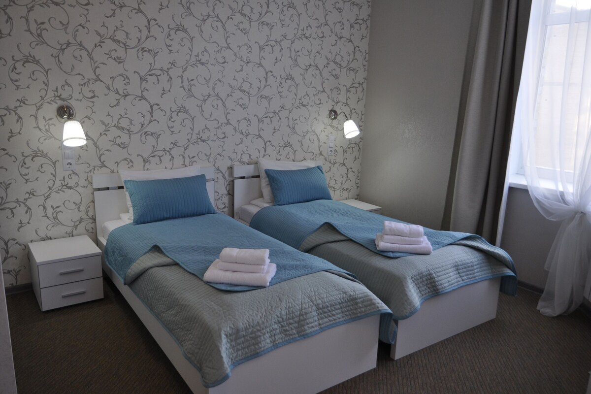 A hotel room with two single beds, each with a blue blanket and white towels. There are two bedside tables with lamps, a window with sheer curtains, and a patterned wallpaper.