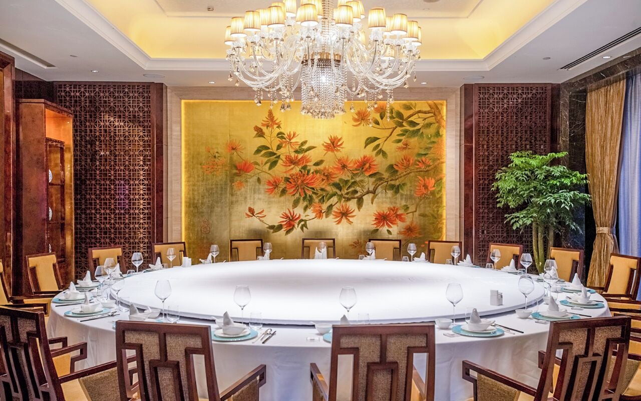 Uma sala de jantar formal com uma mesa redonda posta para refeição, cercada por cadeiras, e um grande candelabro sobre ela.