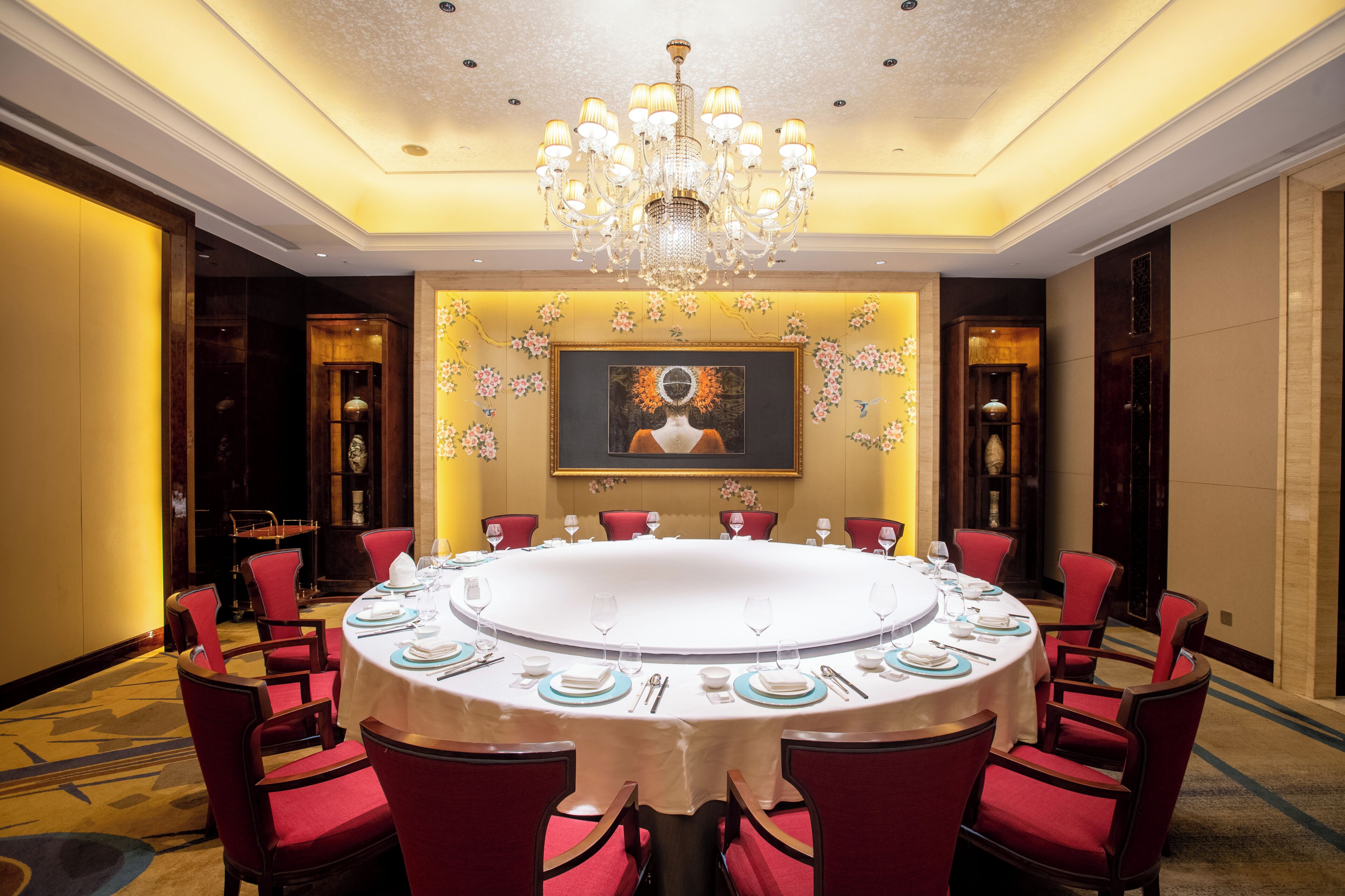 A round table with a white tablecloth, surrounded by red upholstered chairs, in a room with a chandelier and a framed painting on the wall.