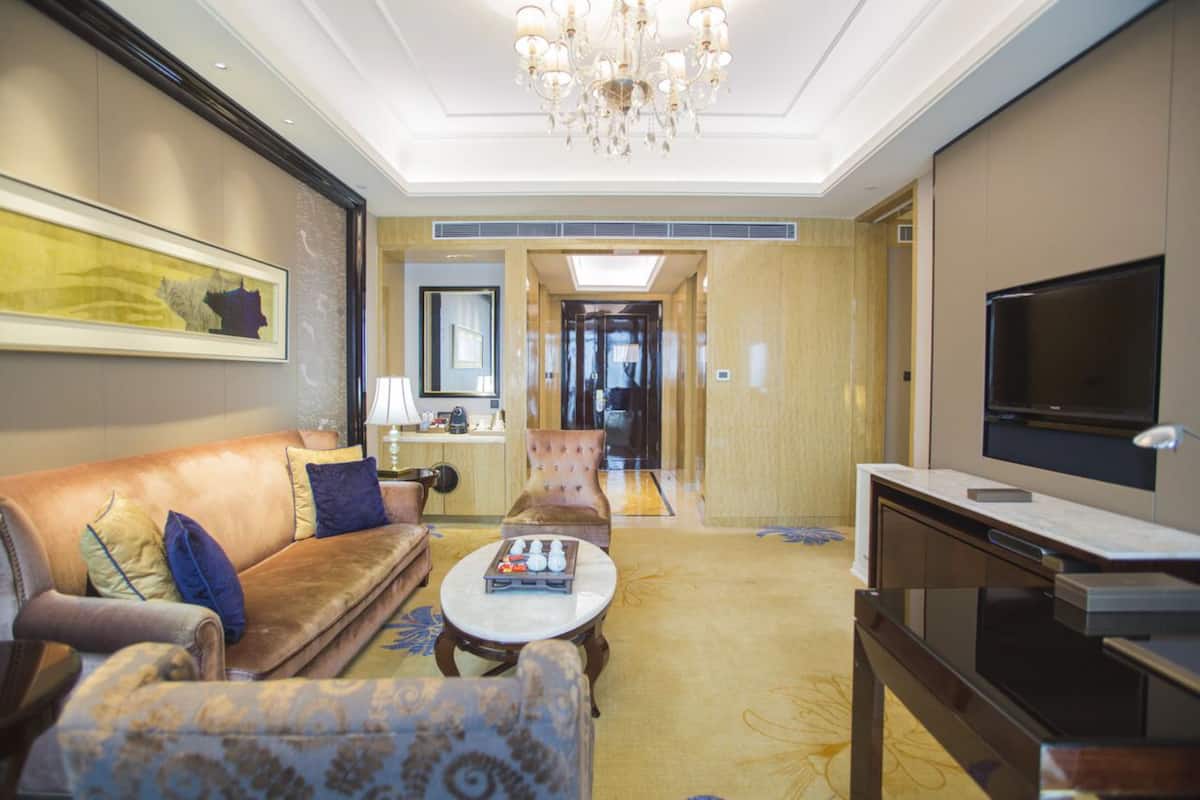 A modern hotel room with a leather sofa, a patterned armchair, a coffee table, a flat-screen TV, and a chandelier.