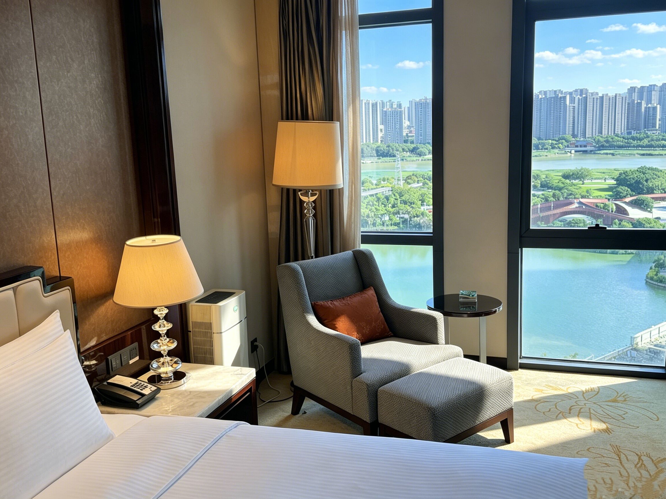 A hotel room with a bed, a chair, a lamp, a small table, and a large window offering a view of a cityscape and a body of water.